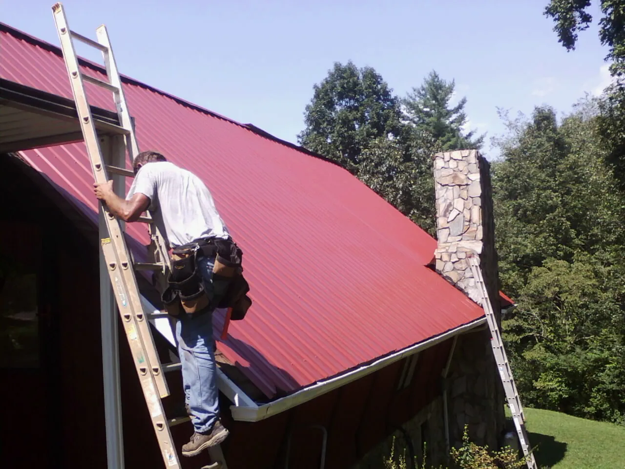 Roofer on a ladder beside a red metal roof and stone chimney — Soffit & Fascia Repair by Roofing Brothers in Savona